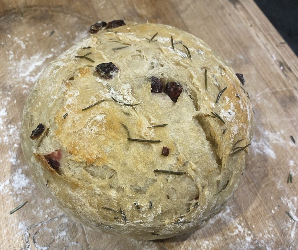 Baked loaf with rosemary leaves and dried cranberry speckling the top resting on a cutting board